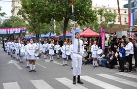 Grupo de jóvenes en uniformes blancos y pantalones cortos, sosteniendo tambores y una bandera, rodeados de espectadores aplaudiendo.