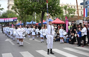 Grupo de jóvenes en uniformes blancos y pantalones cortos, sosteniendo tambores y una bandera, rodeados de espectadores aplaudiendo.