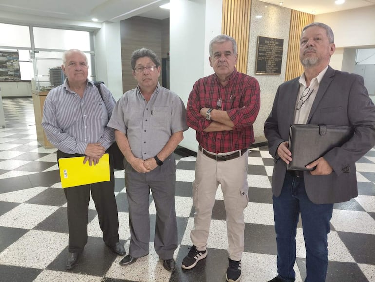 Cuatro hombres en un ambiente interior, con prendas formales y documentos en mano, discuten con calma sobre jubilaciones.