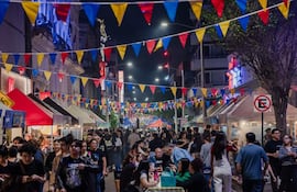 Ambiente festivo con banderas de colores ondeando mientras adultos disfrutan y socializan en puestos de comida durante un evento nocturno.