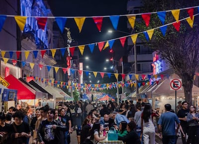 Ambiente festivo con banderas de colores ondeando mientras adultos disfrutan y socializan en puestos de comida durante un evento nocturno.