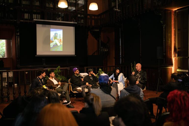 Panelistas en un ambiente acogedor, un hombre con micrófono expone mientras cinco personas lo escuchan atentamente, rodeados de libros.