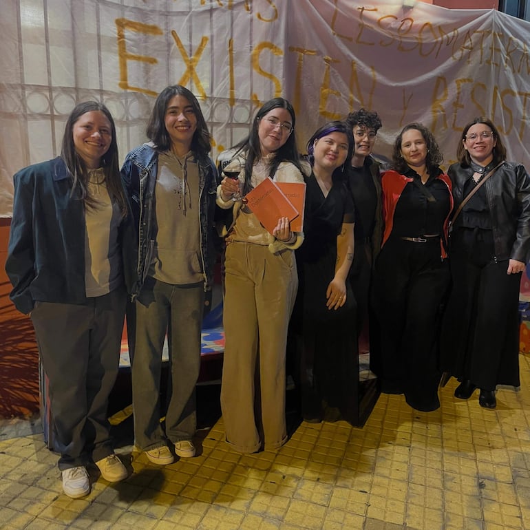 Siete mujeres sonrientes, algunas en ropa formal y otras casual, se agrupan con una carpeta naranjada en un ambiente festivo al aire libre.