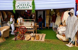 Stand de artesanías en feria al aire libre, con mujeres observando productos de lana en tonos beige y marrones.