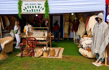 Stand de artesanías en feria al aire libre, con mujeres observando productos de lana en tonos beige y marrones.
