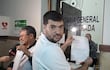 Roberto Riveros sostiene un documento serio, junto a un colega médico con gafas, en un ambiente de oficina con carteles.
