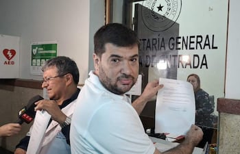Roberto Riveros sostiene un documento serio, junto a un colega médico con gafas, en un ambiente de oficina con carteles.