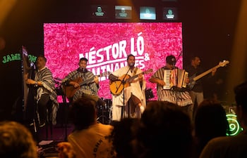 Cinco músicos en el escenario, con vestimentas tradicionales. Tocado el arpa, guitarras y acordeón en un ambiente festivo.