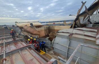 Personas con cascos amarillos y chalecos de salvavidas inspeccionan un barco dañado en un puerto, en un entorno de emergencia.