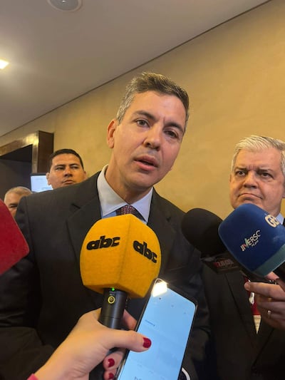 Santiago Peña en traje oscuro, rodeado de micrófonos, mientras Enrique Riera Escudero observa al fondo con cabello canoso.