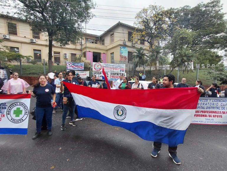 Grupo de manifestantes sostiene carteles y bandera paraguaya, con camisetas de SINALAB, en protesta frente a edificio del MSPyBS.