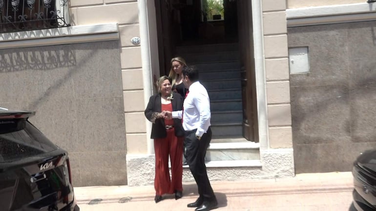 Zenaida Delgado en conjunto cálido con cabello rubio y hombre de camisa blanca y corbata roja, estrechando manos en un entorno urbano.