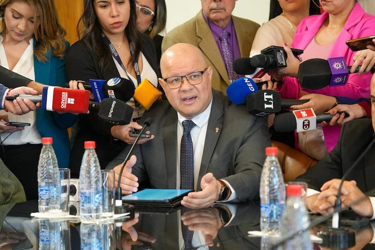 Hombre con traje gris y gafas habla en mesa mientras periodistas sostienen micrófonos, en ambiente formal.