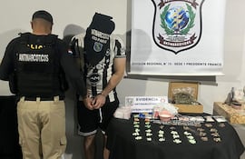 Oficial de policía de espaldas con chaleco antinarcóticos, detenido esposado en camiseta de fútbol, evidencias de drogas sobre la mesa.