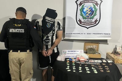 Oficial de policía de espaldas con chaleco antinarcóticos, detenido esposado en camiseta de fútbol, evidencias de drogas sobre la mesa.