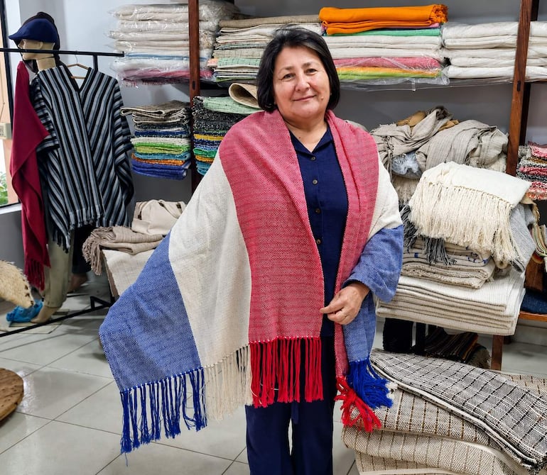 Julia Álvarez sonríe sosteniendo un poncho rojo, blanco y azul en una tienda de textiles con mantas de diversos colores.