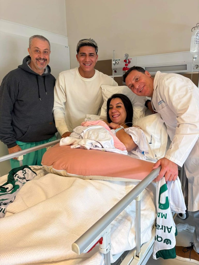 Mujer en cama sonríe con su bebé, rodeada de tres hombres; uno con barba, otro de cabello claro y un médico en bata.
