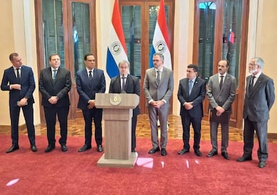 Hombres de pie en traje oscuro, atentos al orador en el podio con banderas de Paraguay en el fondo.