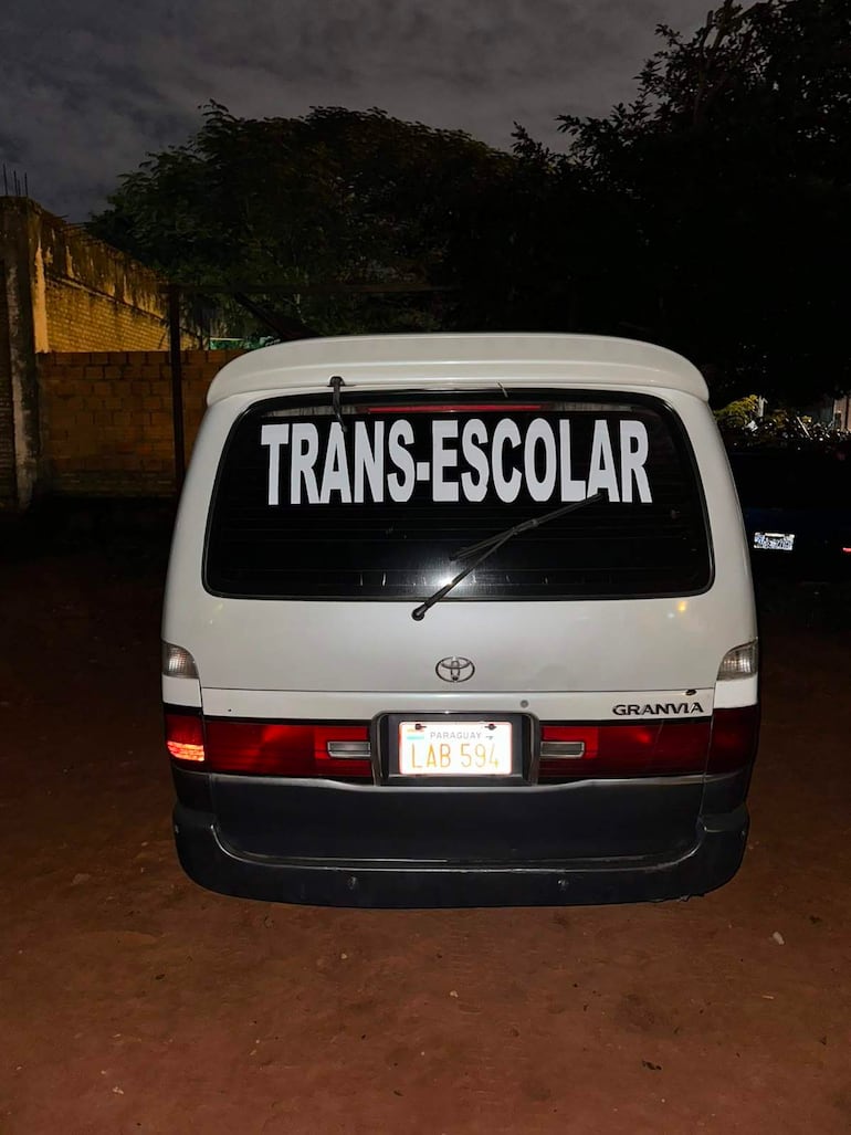 Furgoneta blanca con detalles rojos y letrero 'TRANS-ESCOLAR' visible en el ventanal trasero, estacionada en un área de tierra.