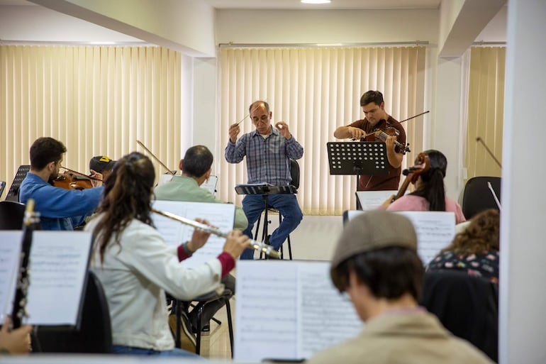 Gabriel Graziani sentado dirigiendo con batuta, violinista concentrado a su izquierda, instrumentos visibles en primer plano.