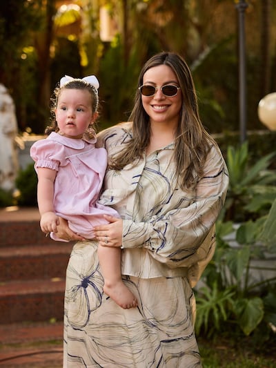 Mujer con gafas de sol y blusa abstracta sostiene a niña en vestido rosa con lazo blanco, ambas sonriendo en un entorno verde.