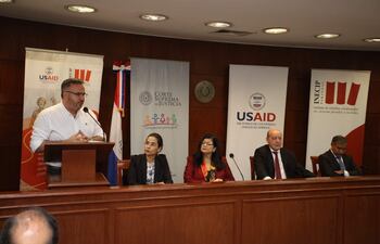 Hombre con camisa blanca habla en un micrófono; mujeres y hombres escuchan atentamente rodeados de banners de USAID y la Corte Suprema.
