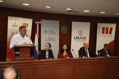 Hombre con camisa blanca habla en un micrófono; mujeres y hombres escuchan atentamente rodeados de banners de USAID y la Corte Suprema.