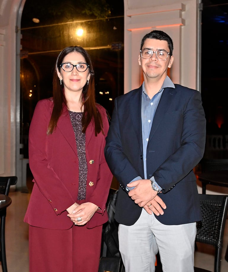 Mujer de cabello largo y lacio con atuendo burdeos sonríe junto a hombre con blazer oscuro y gafas, en un elegante salón.