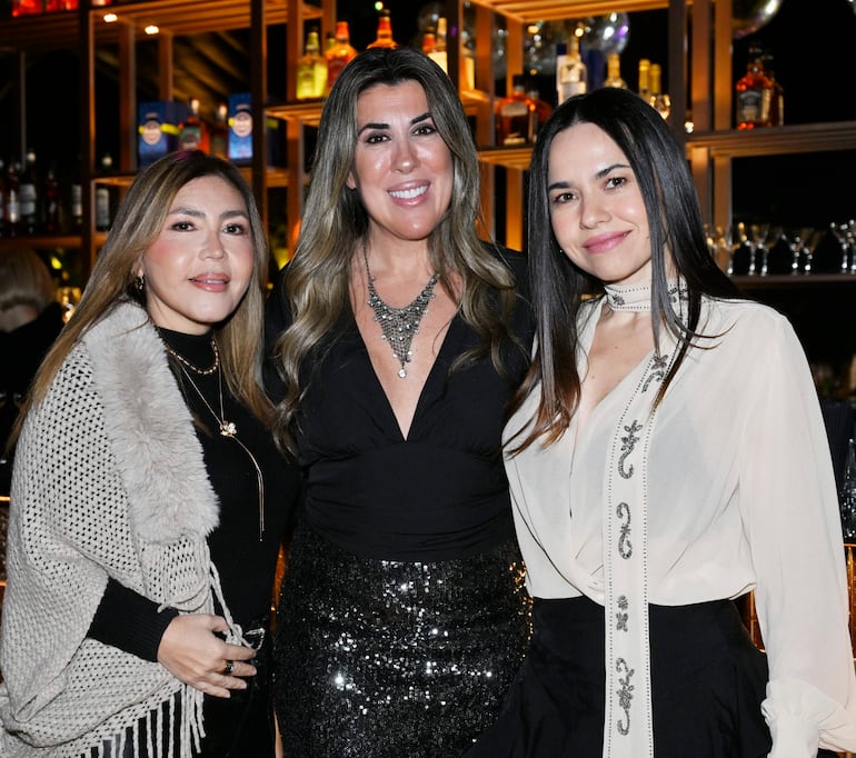 Tres mujeres sonrientes en un bar: una con vestido negro, otra con suéter gris claro y otra con blusa blanca decorada.
