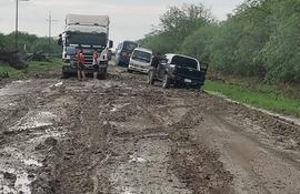 Dos hombres en camisetas rojas junto a un camión blanco en una carretera llena de barro, rodeados de vegetación.