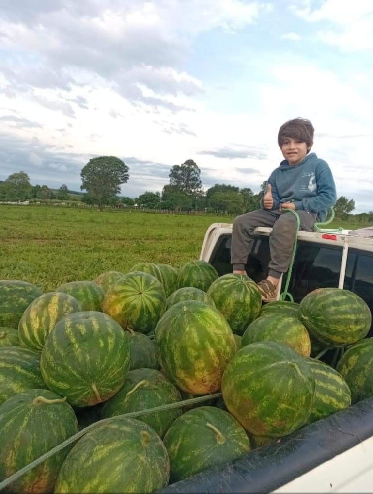 Niño de cabello castaño claro con abrigo azul, sonríe mostrando el pulgar hacia arriba en camioneta con sandías en un campo verde.