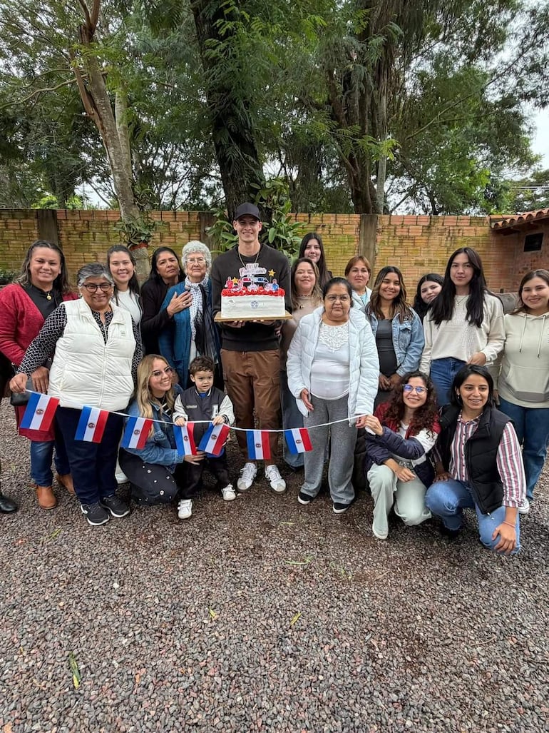 Hombre alto sostiene un pastel decorado rodeado de personas sonrientes con banderas pequeñas de Paraguay en un ambiente natural.