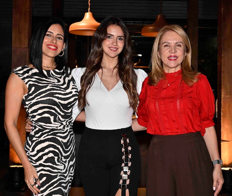 Tres mujeres sonrientes: una en blusa blanca y pantalón negro, otra en blusa roja y falda oscura, en un ambiente bien iluminado.