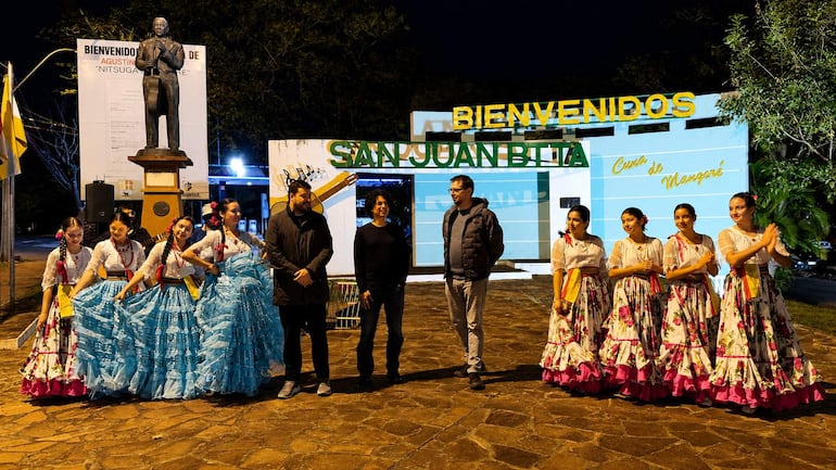Tres hombres en ropa casual conversan, rodeados de bailarinas en trajes tradicionales, sonriendo en un evento nocturno.