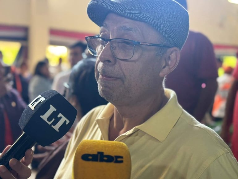 Hombre mayor en primer plano con gorra y gafas, sostiene un micrófono rodeado de periodistas en un ambiente público.