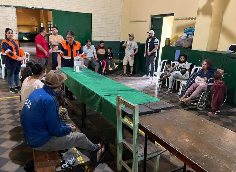 Ocho personas sentadas alrededor de una mesa verde, con botellas de agua, en un ambiente de asistencia social. Dos llevan chaquetas naranjas.