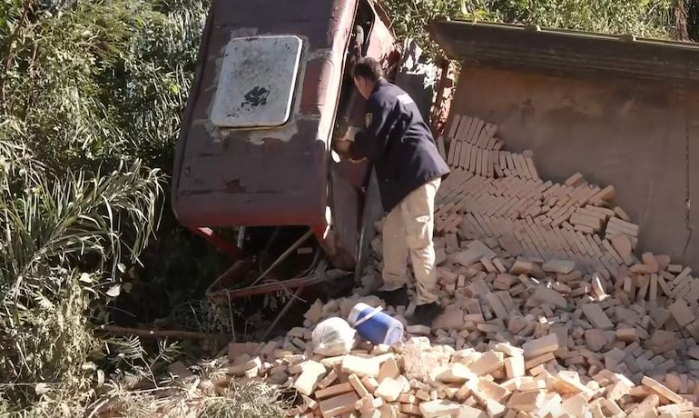 Hombre con abrigo oscuro y pantalones claros frente a camión volcado lleno de ladrillos, rodeado de vegetación.
