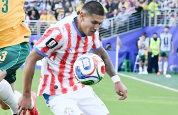Juan Cáceres en camiseta roja y blanca con balón, frente a un jugador australiano en camiseta amarilla, en un estadio lleno.