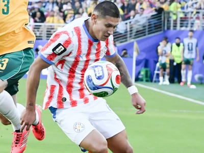 Juan Cáceres en camiseta roja y blanca con balón, frente a un jugador australiano en camiseta amarilla, en un estadio lleno.