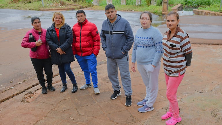 Grupo de seis adultos en calle de tierra, dos con chaquetas, una mujer con suéter gris y otra con camiseta de rayas, posando juntos.
