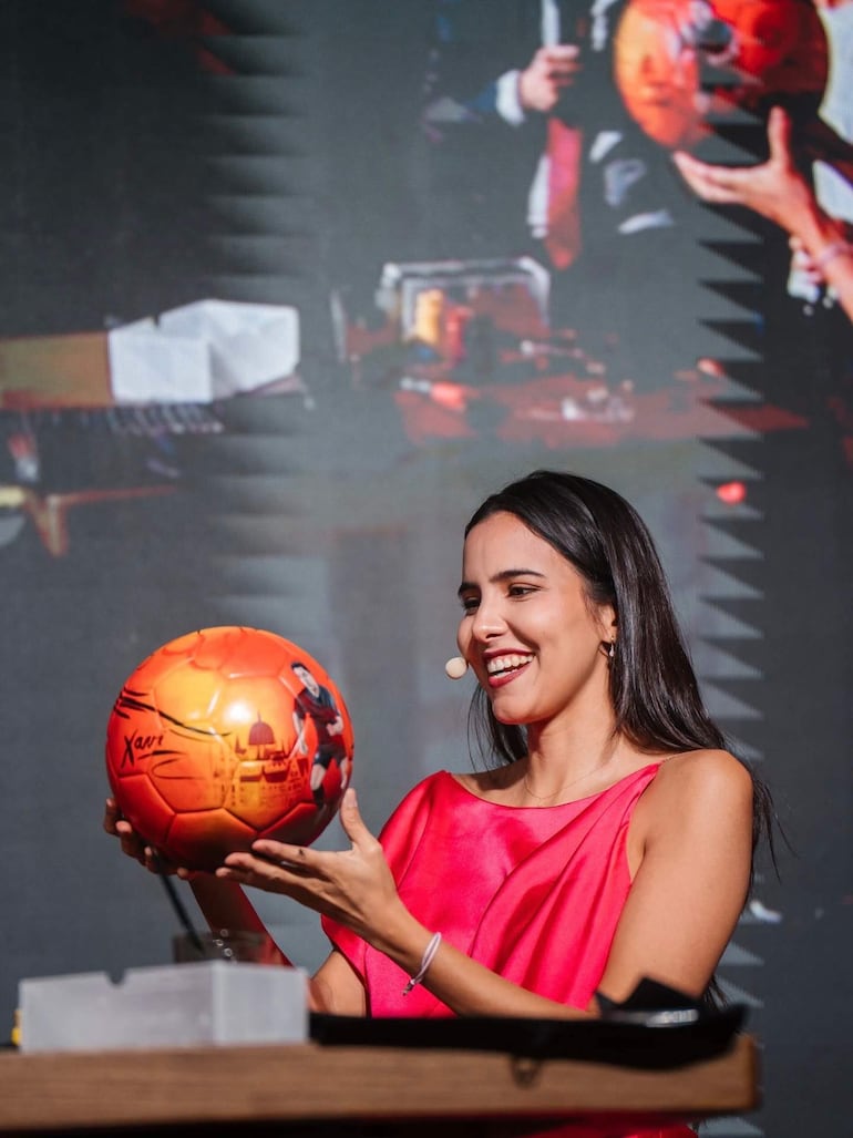 Mujer con blusa rosa sonríe sosteniendo un balón con diseño de jugador, mientras un hombre la acompaña en segundo plano.