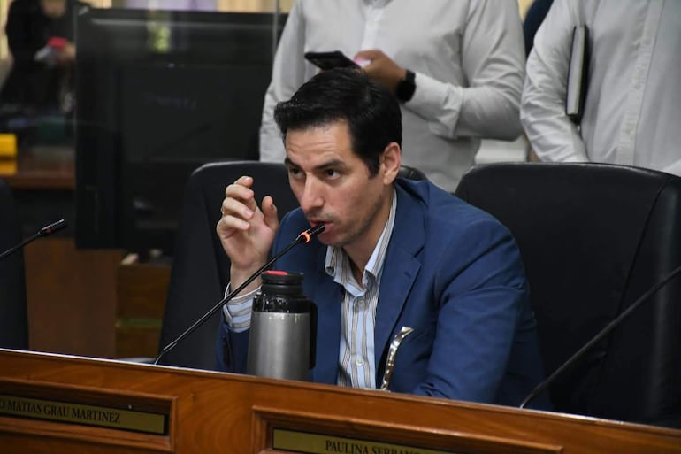 Álvaro Grau en chaqueta azul y camisa blanca, sosteniendo un objeto, durante un debate en un entorno formal con personas observando.