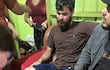 Almir de Brum sentado con expresión seria en sofá, acompañado de un hombre y una mujer que lo asisten en un ambiente doméstico.