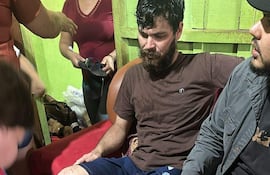Almir de Brum sentado con expresión seria en sofá, acompañado de un hombre y una mujer que lo asisten en un ambiente doméstico.