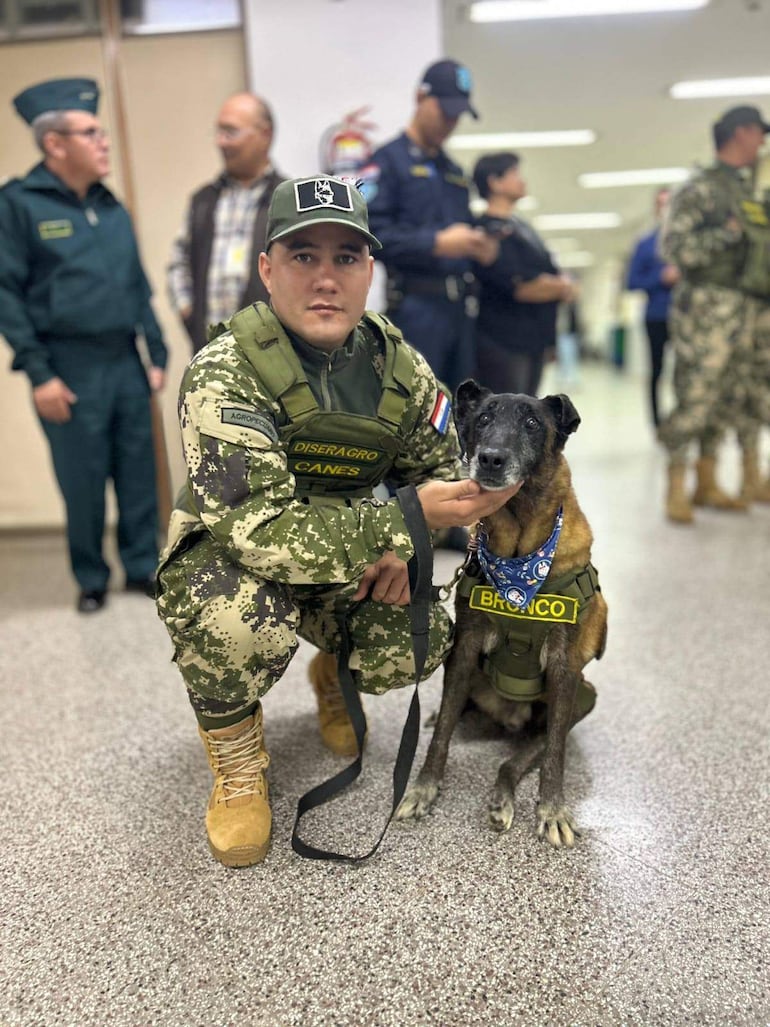 Sgto Gustavo Gómez en uniforme militar acaricia al perro Bronco en ceremonia institucional con otros asistentes de fondo.