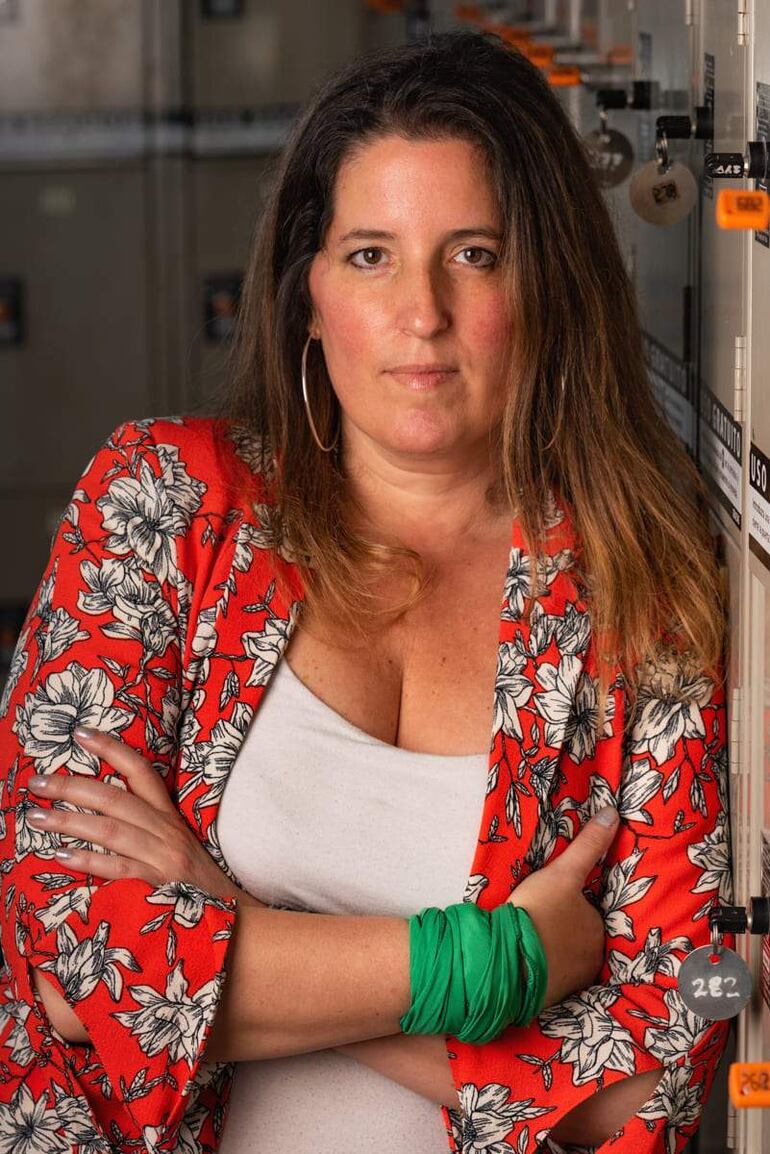 Mujer con chaqueta roja y camiseta blanca, cruzando los brazos con confianza, en un entorno de trabajo con armarios al fondo.