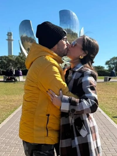 Hombre con abrigo amarillo y gorro negro besa a mujer con abrigo de cuadros, en un parque con escultura moderna de fondo.