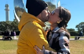 Hombre con abrigo amarillo y gorro negro besa a mujer con abrigo de cuadros, en un parque con escultura moderna de fondo.