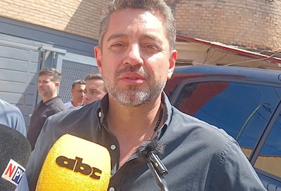 Pedro Alliana con camisa oscura sostiene un micrófono mientras un grupo de hombres lo observa en el fondo, ambiente exterior con pared de ladrillos.