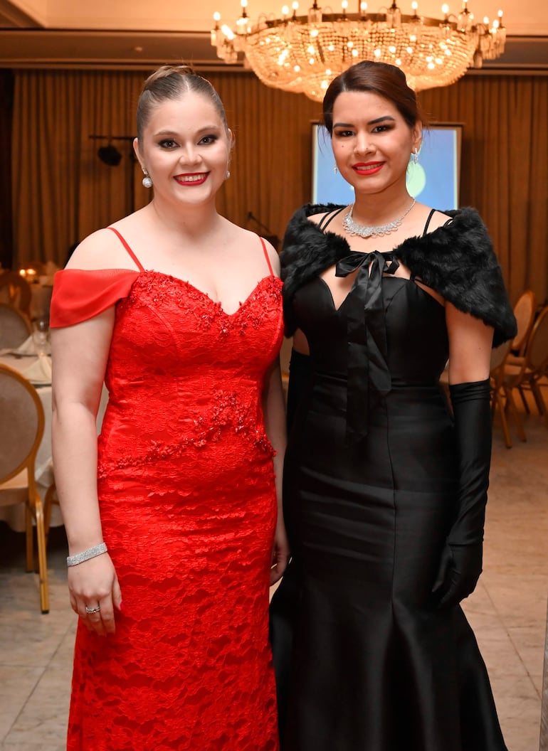 Mujeres sonrientes en gala, una con vestido rojo y otra con vestido negro, en un salón elegantemente decorado.